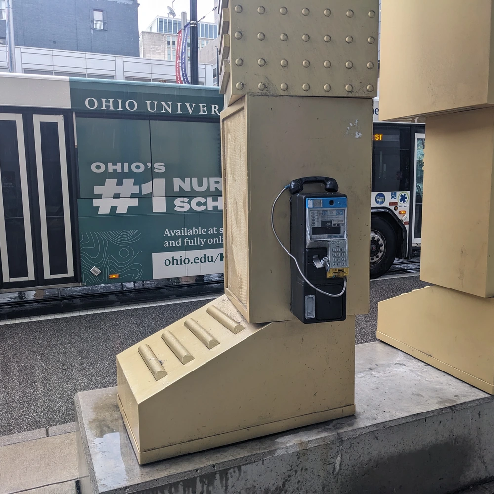 Beige steel base of a sculpture with a pay phone mounted on its front and two plaques on the columns, on a city sidewalk.