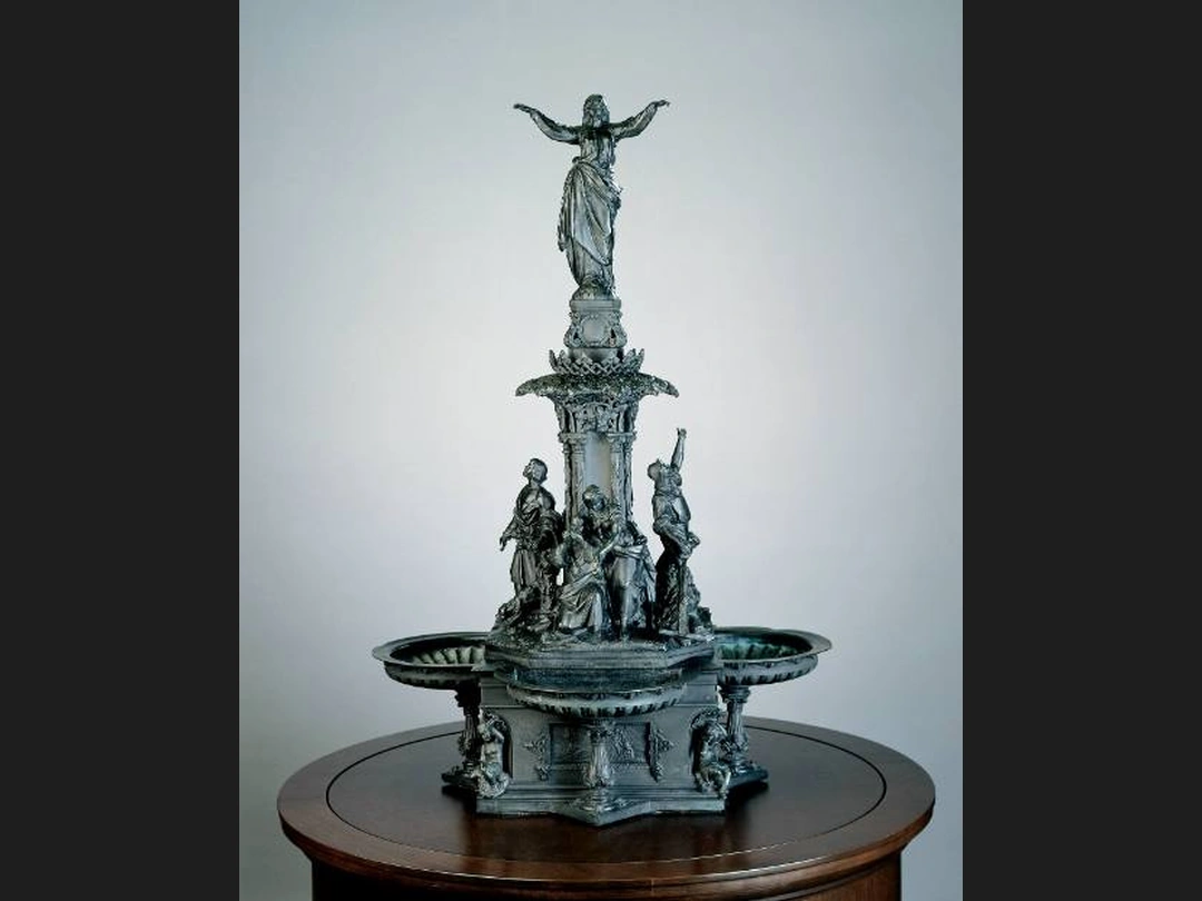 Bronze model of a tiered fountain, with a standing figure with outstretched arms at the top and figure groups around the central column, on a round wooden pedestal against a grey background.