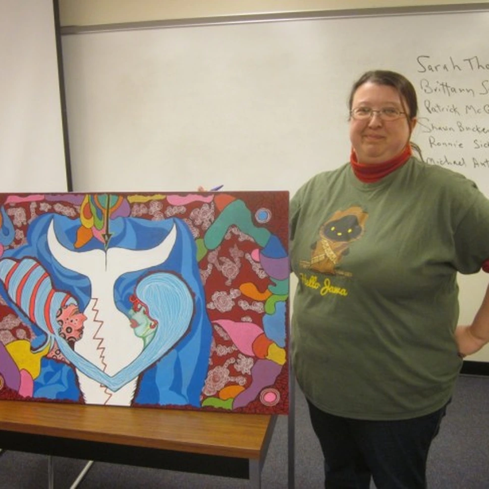 A smiling woman in a green T-shirt stands beside her painting of two abstract figures in blue and white, in a classroom with a whiteboard.