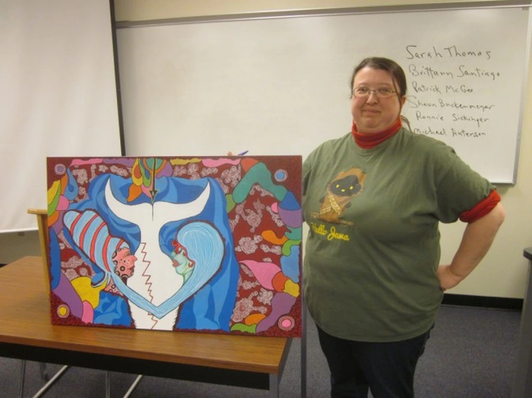 A smiling woman in a green T-shirt stands beside her painting of two abstract figures in blue and white, in a classroom with a whiteboard.