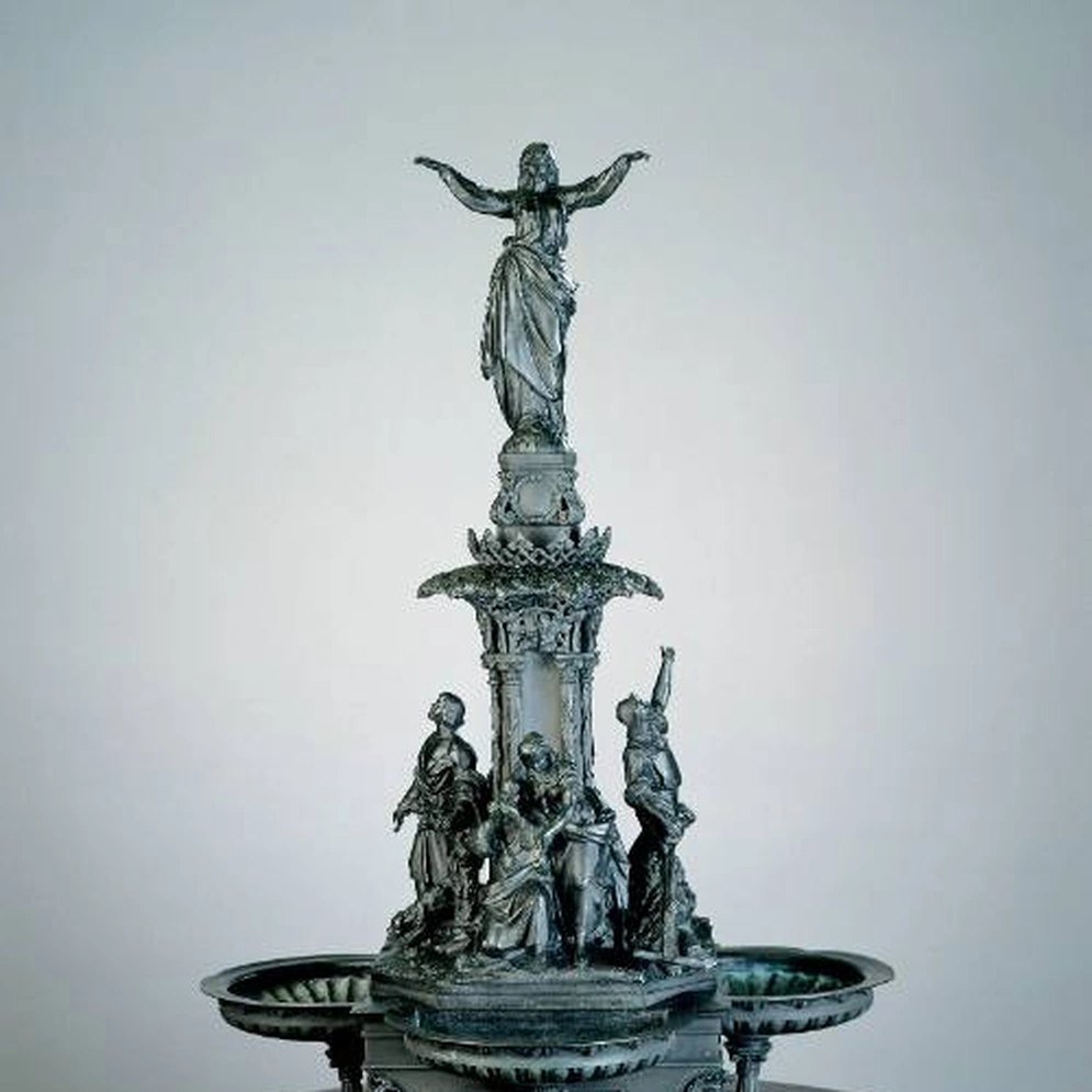Bronze model of a tiered fountain, with a standing figure with outstretched arms at the top and figure groups around the central column, on a round wooden pedestal against a grey background.