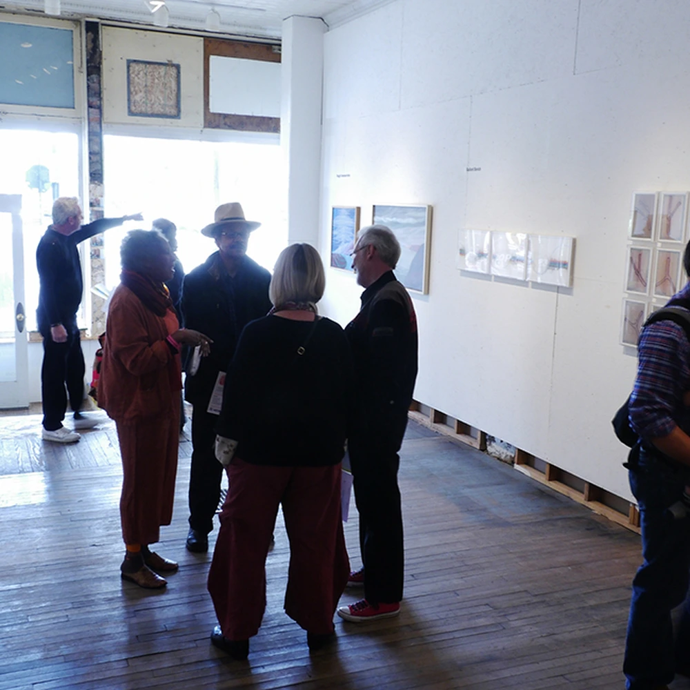 A group of visitors in a bare-floored gallery room with white walls, standing and talking near small framed prints, one man pointing.