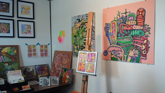Studio wall with colorful painted canvases and framed small works above a table of prints and objects.