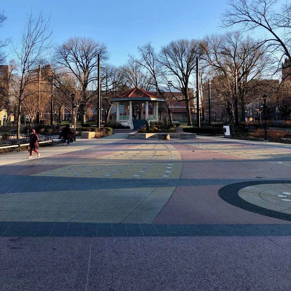 Red-roofed bandstand pavilion in a paved park plaza, with bare trees under a clear blue sky.