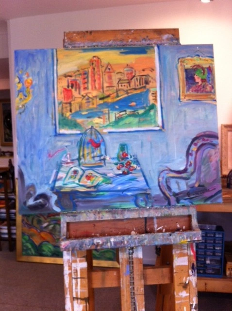 Easel holding a blue painting of an interior with a birdcage, a bowl of flowers and a small city skyline above.