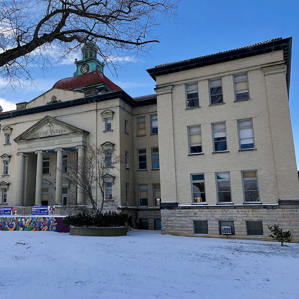 Columned portico and clock tower of the old Clifton school building, seen past a bare tree in the snow.