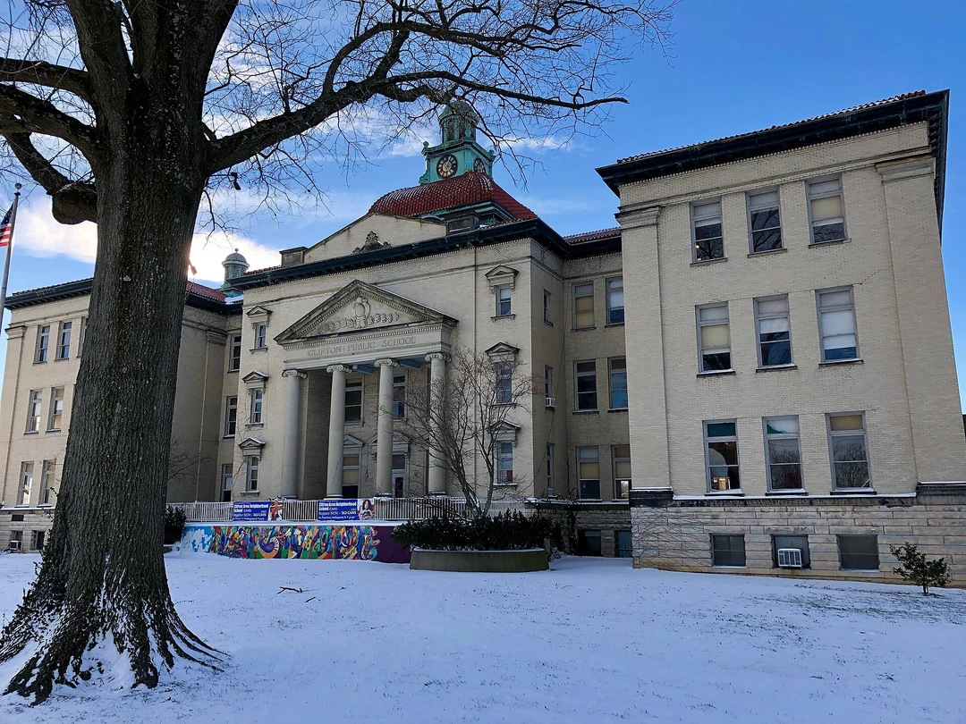 Brick school building with a columned portico, a copper clock tower and a red roof, behind a bare tree in the snow.