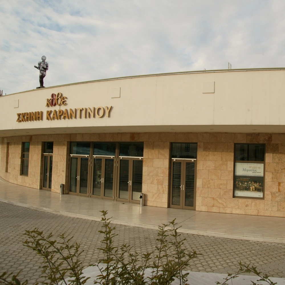 Two-storey cream stone building with a curved roof, glass doors and the sign Σκηνή Καραντίνου.