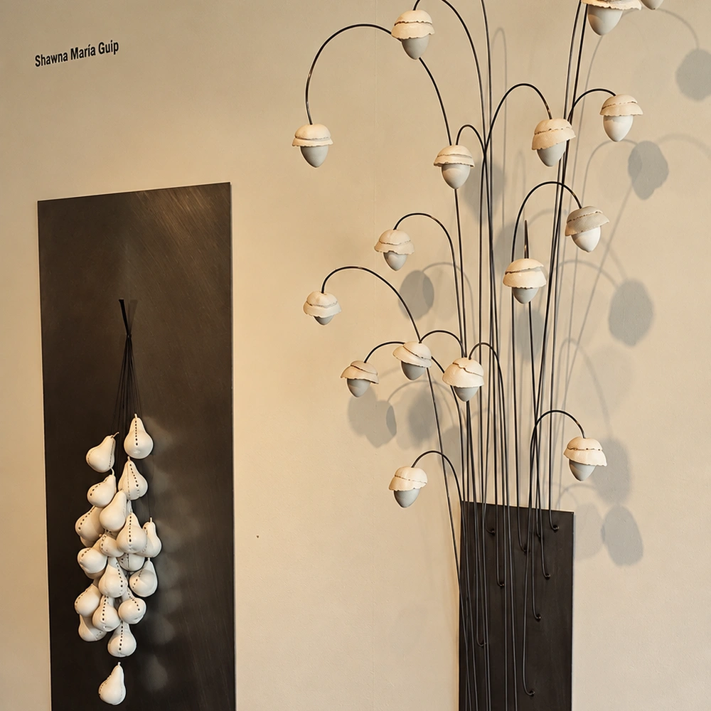 Thin steel rods with pale porcelain blossoms rising from a dark steel panel on a cream wall.