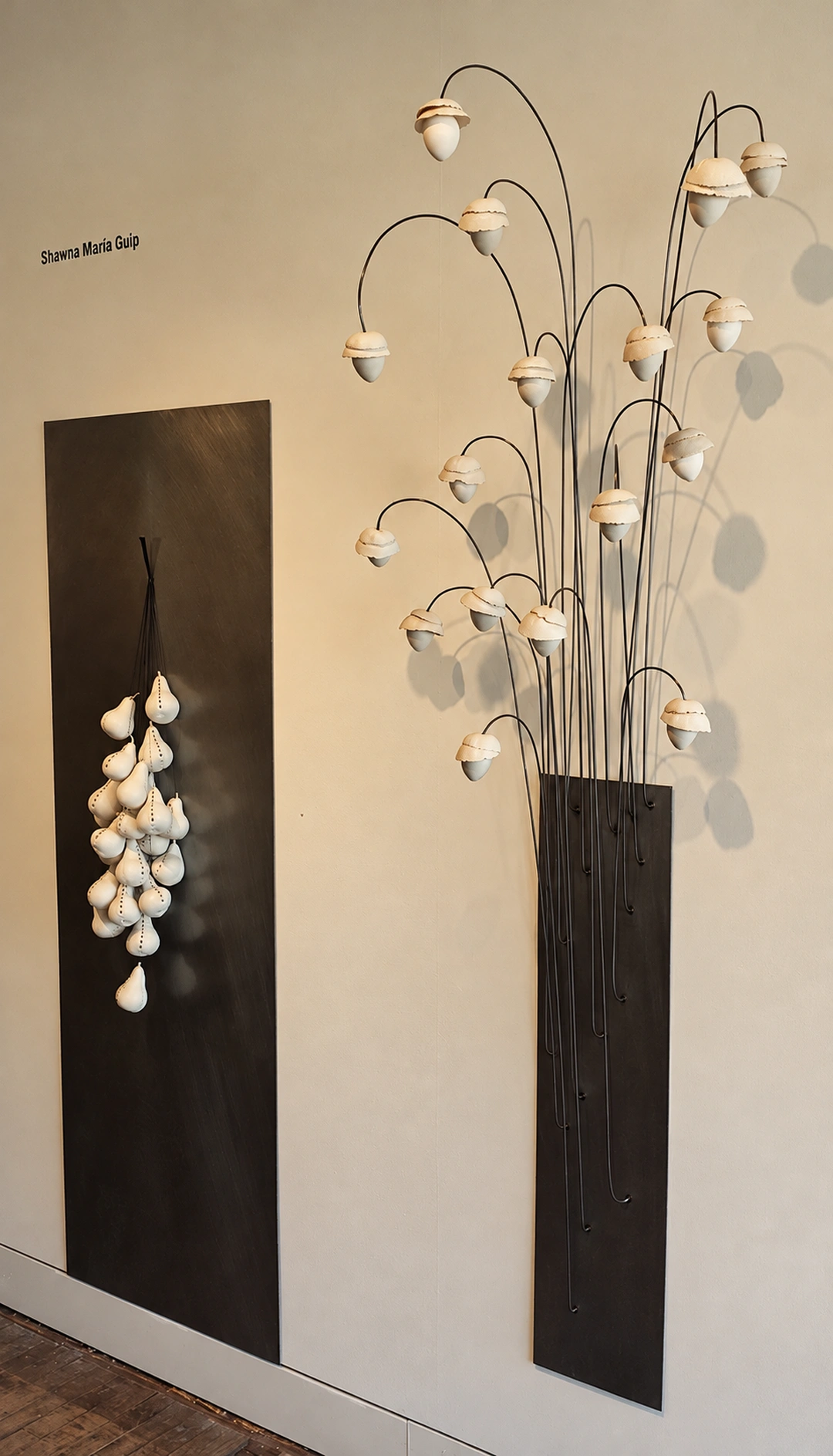 Two dark steel wall panels with white porcelain forms: a cluster hangs on the left, and tall thin rods with pale cup-shaped blossoms rise on the right.