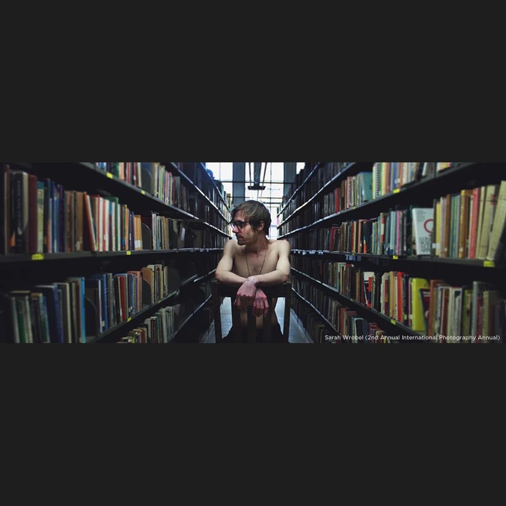 Photograph of a shirtless man with glasses seated on a chair between rows of bookshelves in a library.
