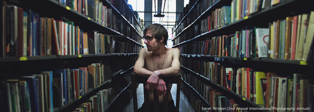 Photograph of a shirtless man with glasses seated on a chair between rows of bookshelves in a library.