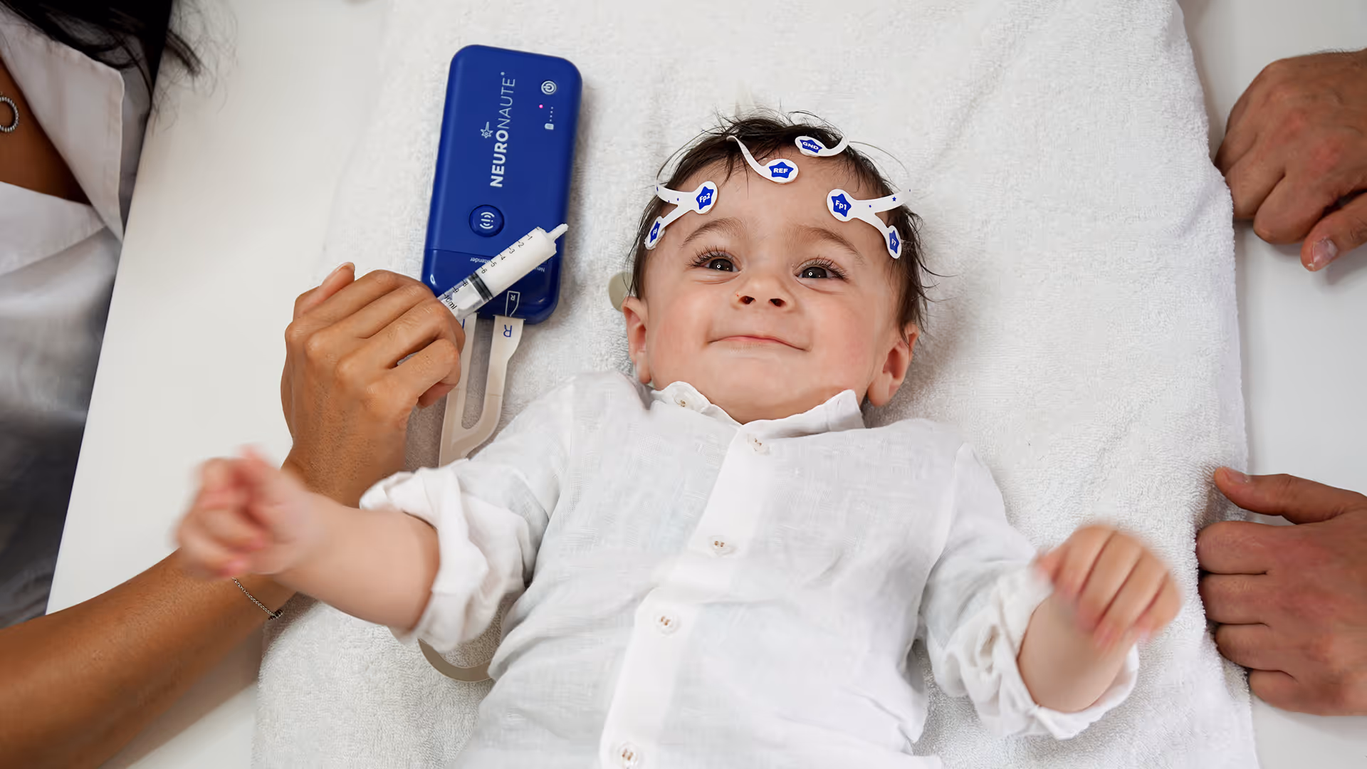 Smiling baby lying on a white towel with EEG sensors attached to the head and a person holding a medical device and syringe nearby.