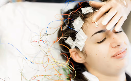 Young woman with electrodes and wires attached to her head, lying down with eyes closed during a sleep study or brain monitoring.