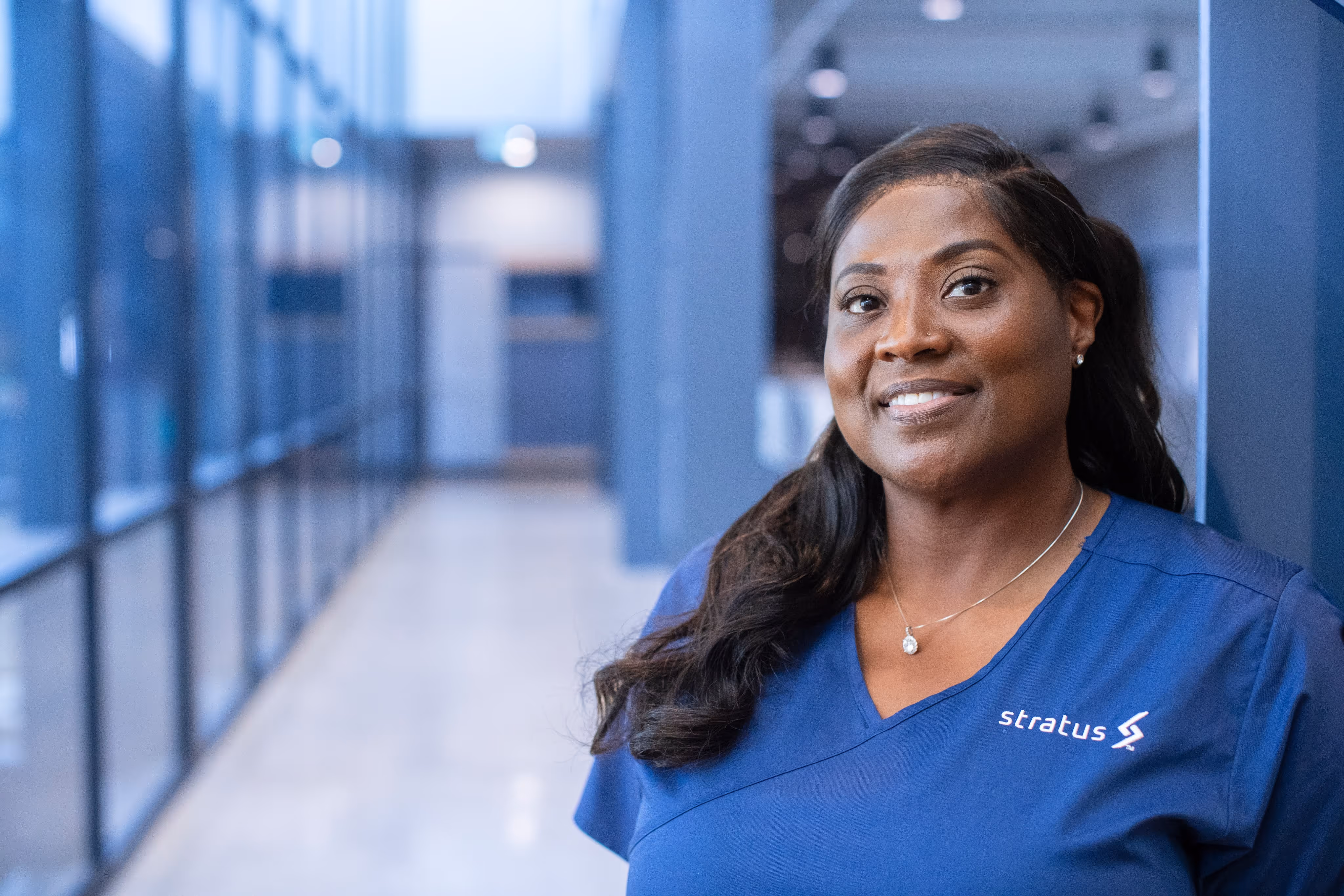 Smiling woman in blue scrubs with a Stratus logo standing in a bright modern hallway.