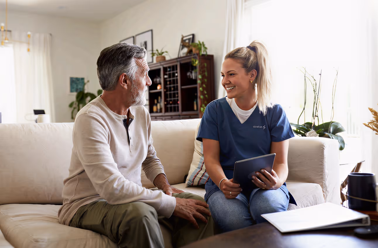 A female healthcare worker in blue scrubs smiling and holding a tablet while talking to an older man sitting on a couch.