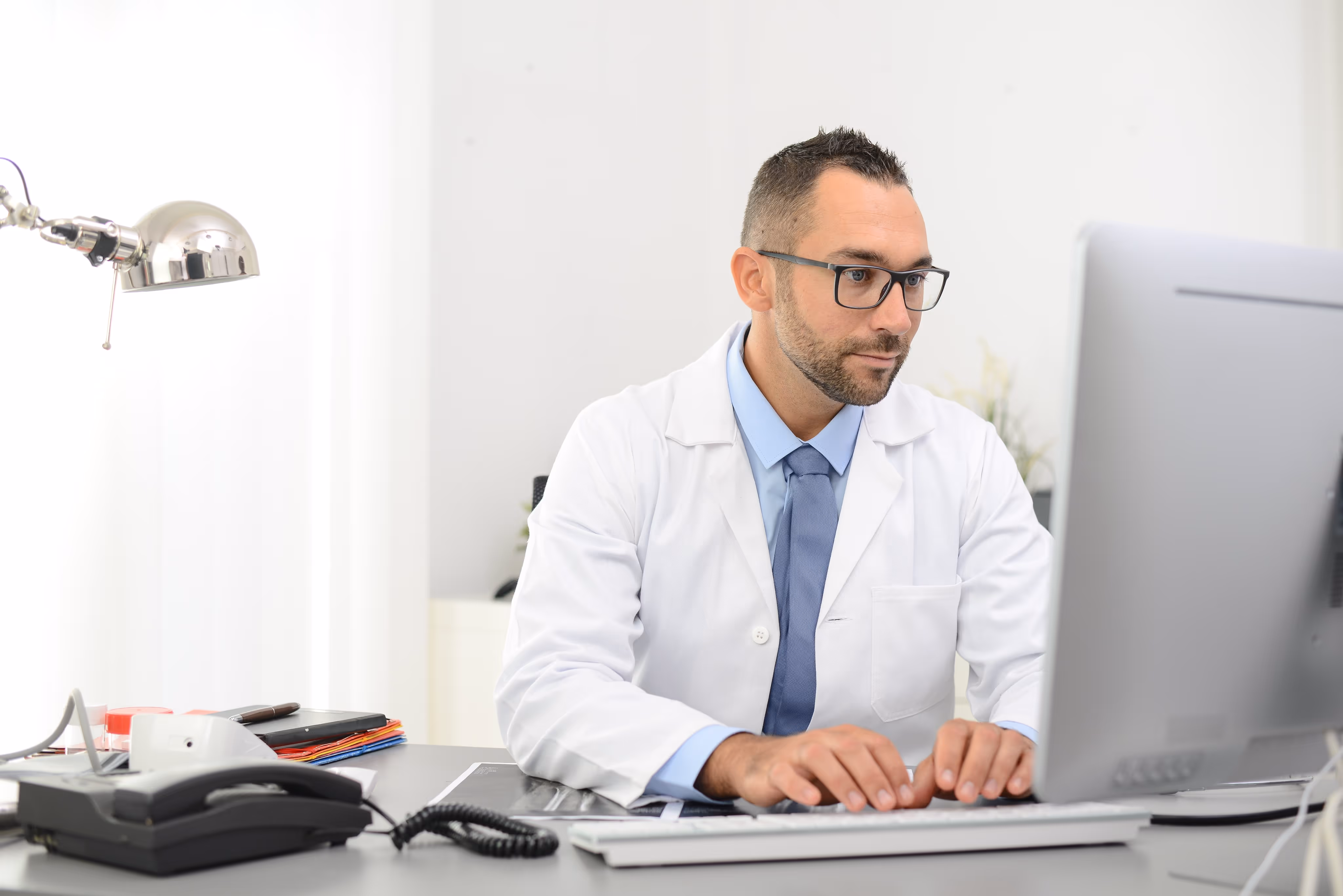 Male doctor in a white coat and glasses typing on a keyboard at a desk with a computer and office phone.