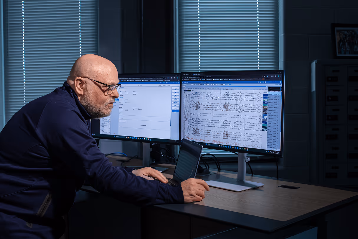 Bald man with glasses working on a laptop at a desk with two large monitors displaying data and waveforms.