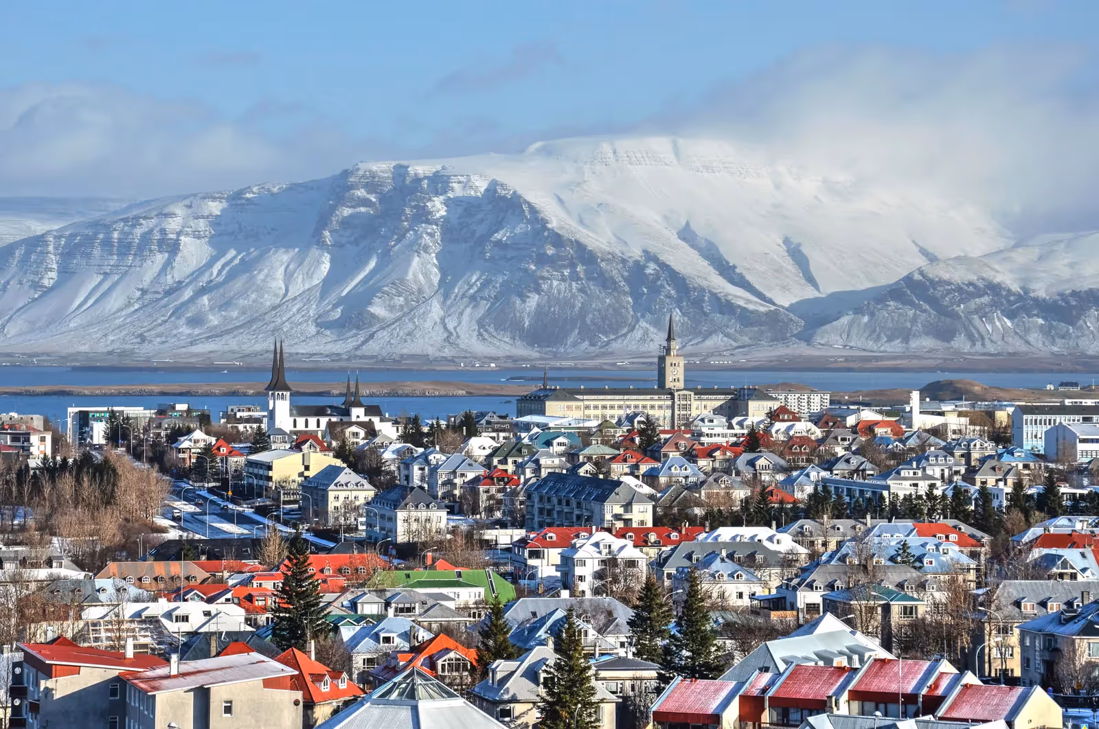 View of a snowy cityscape with colorful rooftops, two church steeples, and a large snow-covered mountain in the background.