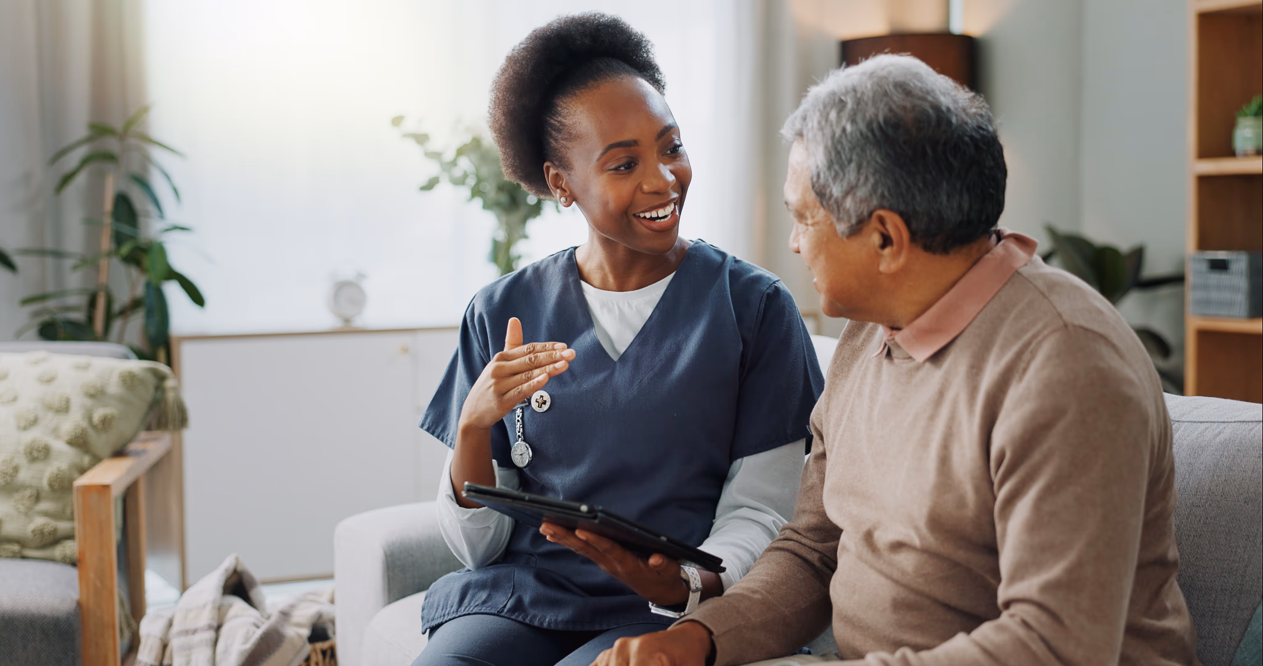 A smiling nurse in blue scrubs talks with an elderly man on a couch in a cozy living room.