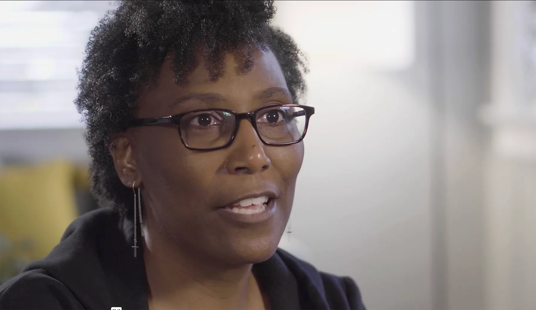Close-up of a woman with short curly hair, wearing glasses and cross earrings, speaking indoors.