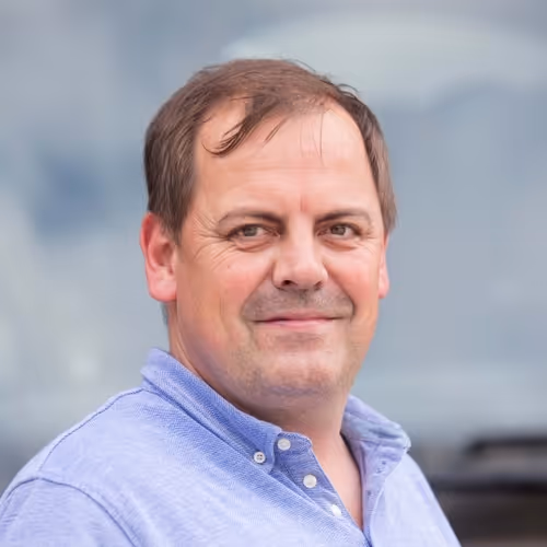 Middle-aged man with short brown hair wearing a light blue button-up shirt smiling softly, with a blurred outdoor background.