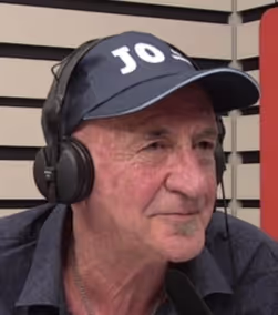 Older man wearing a navy cap with white lettering, headphones, and a dark shirt, looking to the side.