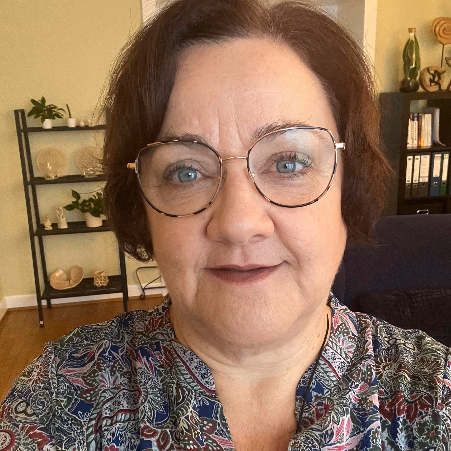 Middle-aged woman with short brown hair, wearing large round glasses and a patterned blouse in a room with shelves and books.