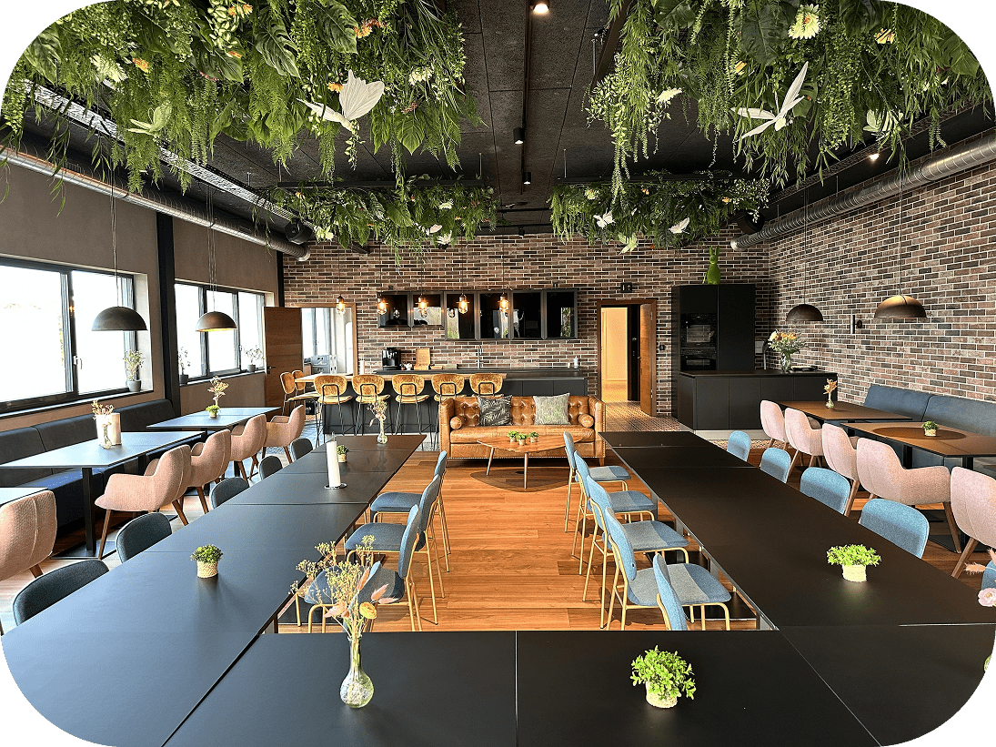Modern conference room with black tables arranged in a U shape, blue and pink chairs, hanging greenery with white butterflies, exposed brick walls, and a lounge area with a brown leather sofa.