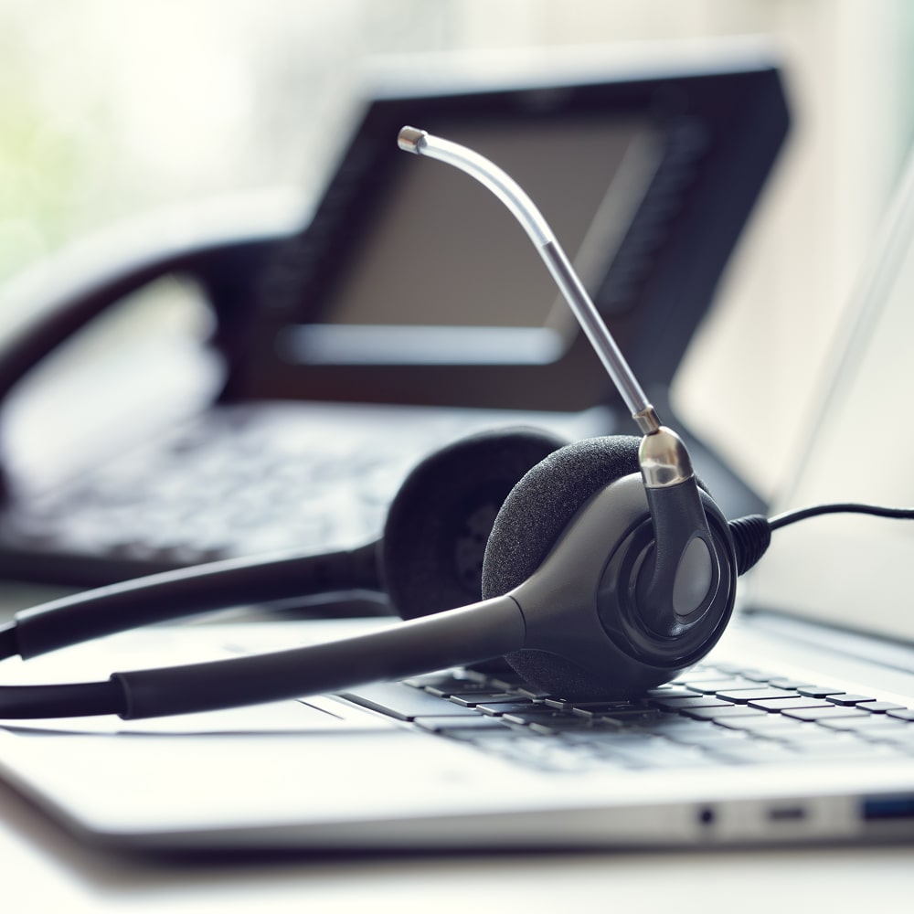 Black headset with microphone resting on a laptop keyboard with a blurred office phone in the background.
