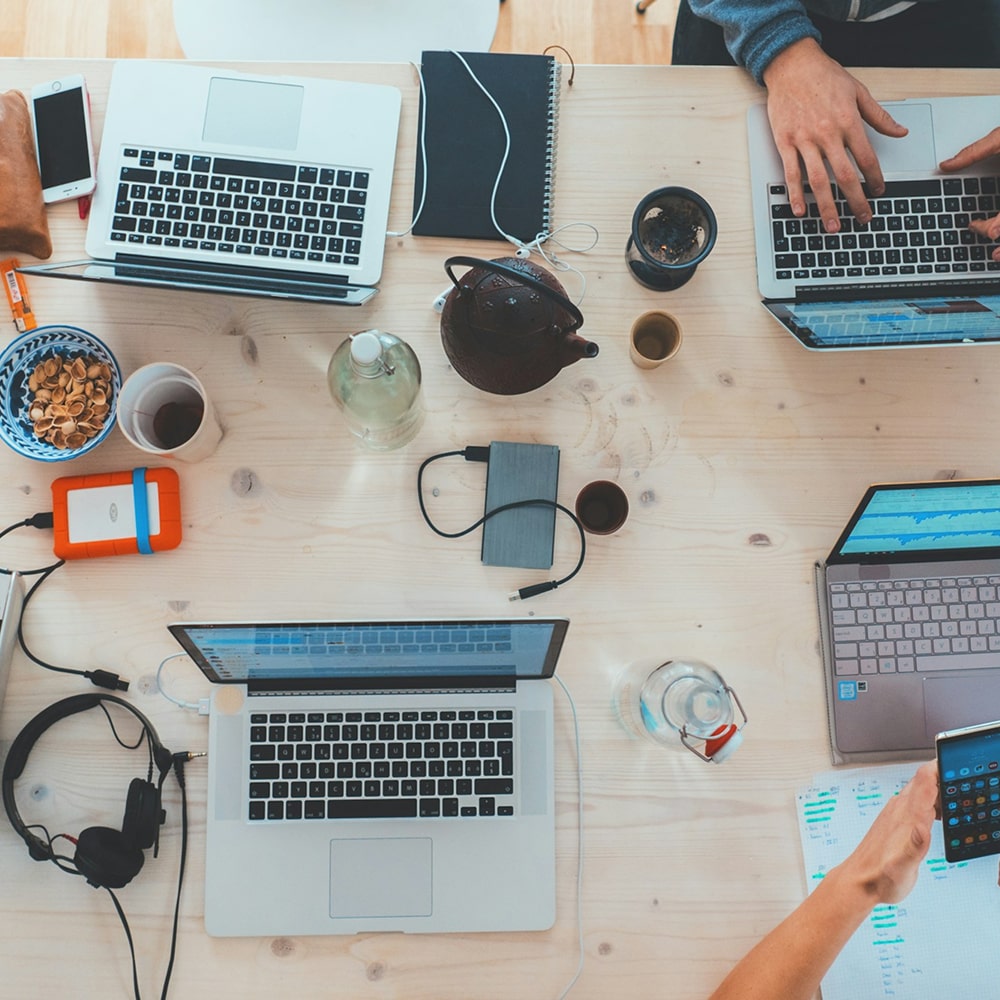 Top-down view of a wooden table with three laptops, headphones, notebooks, tea set, water bottles, and hands using a laptop and smartphone.