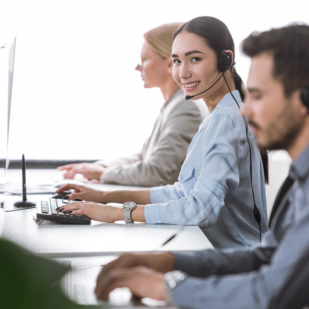 Smiling woman wearing headset and typing on keyboard in a busy call center with colleagues.