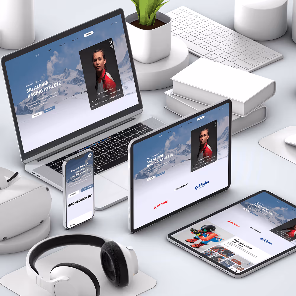 Multiple devices including a laptop, tablet, and smartphone displaying a ski alpine racing athlete website with sponsor logos, surrounded by headphones, books, and a keyboard on a white desk.