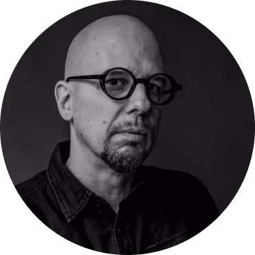 Black and white portrait of a bald man with glasses and a short beard wearing a dark shirt, against a dark background.