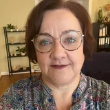 Middle-aged woman with short brown hair, wearing large round glasses and a patterned blouse in a room with shelves and books.