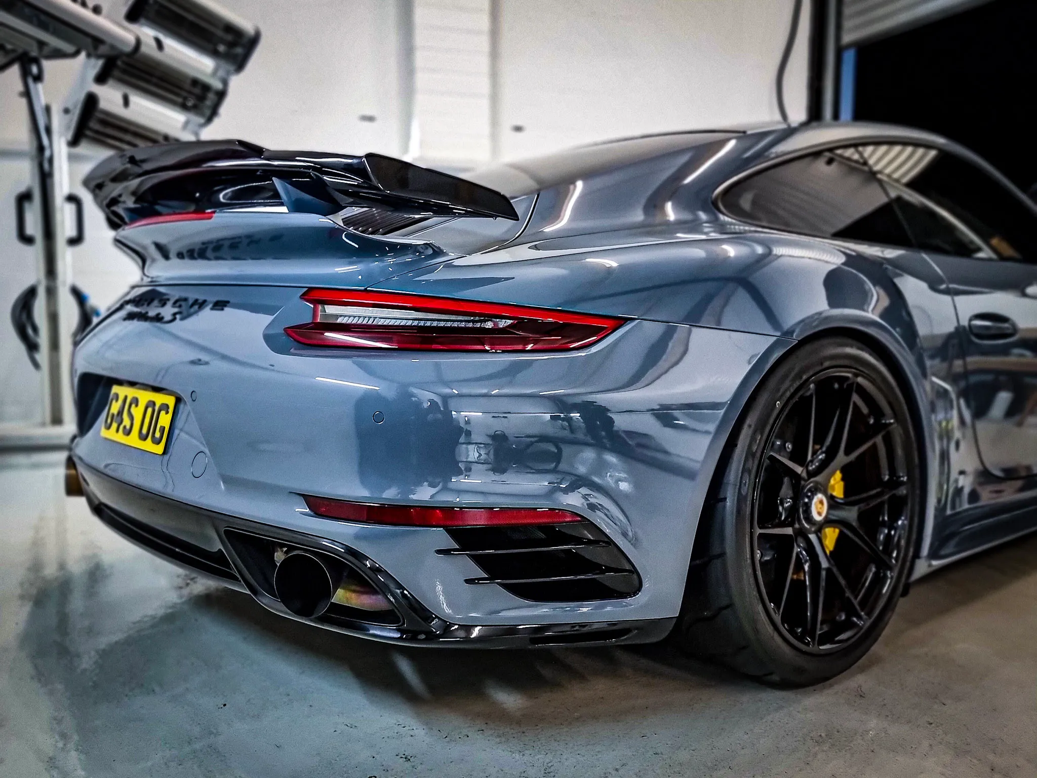 Rear view of a gray Porsche 911 Turbo S with black rims and yellow brake calipers inside a garage.