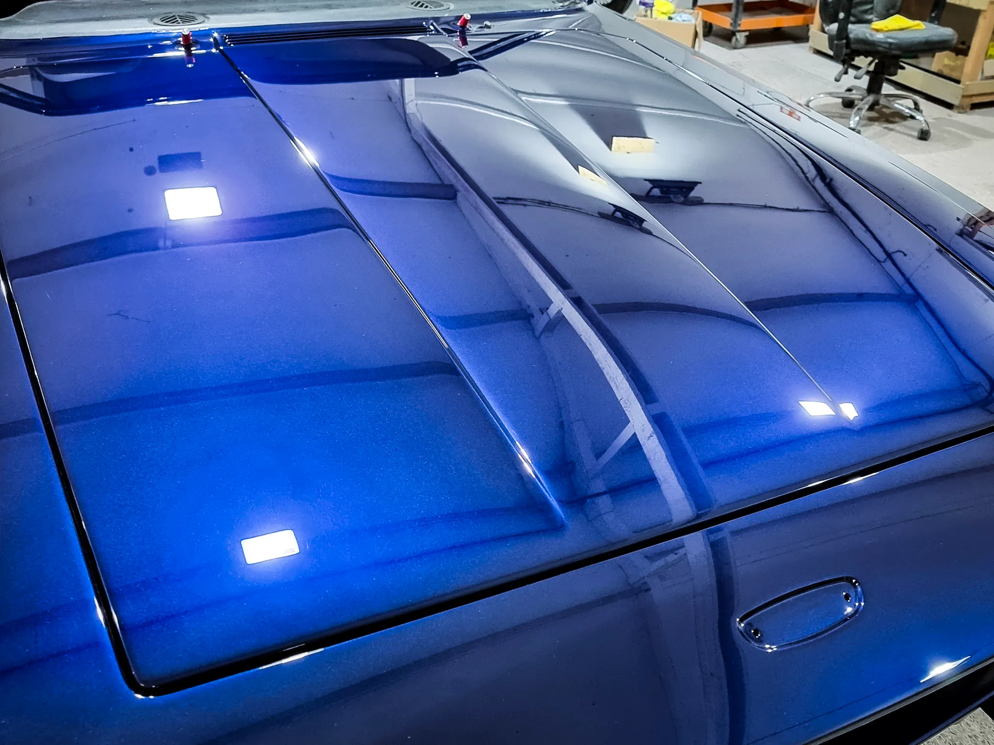 Glossy blue car hood with raised air intake vents reflecting indoor ceiling lights and workshop surroundings.