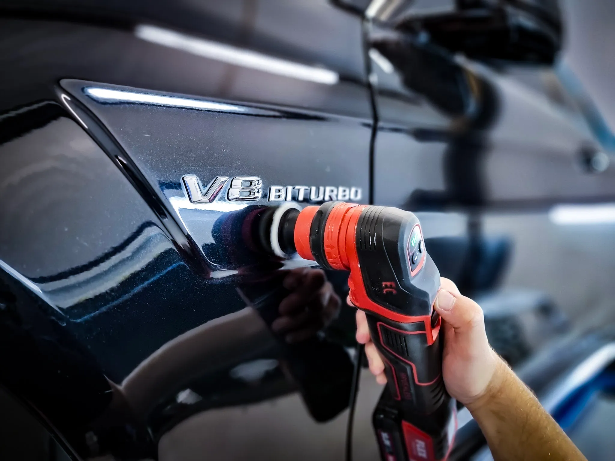 A hand holding a red and black polishing machine buffing the side of a black car near a V8 Biturbo emblem.