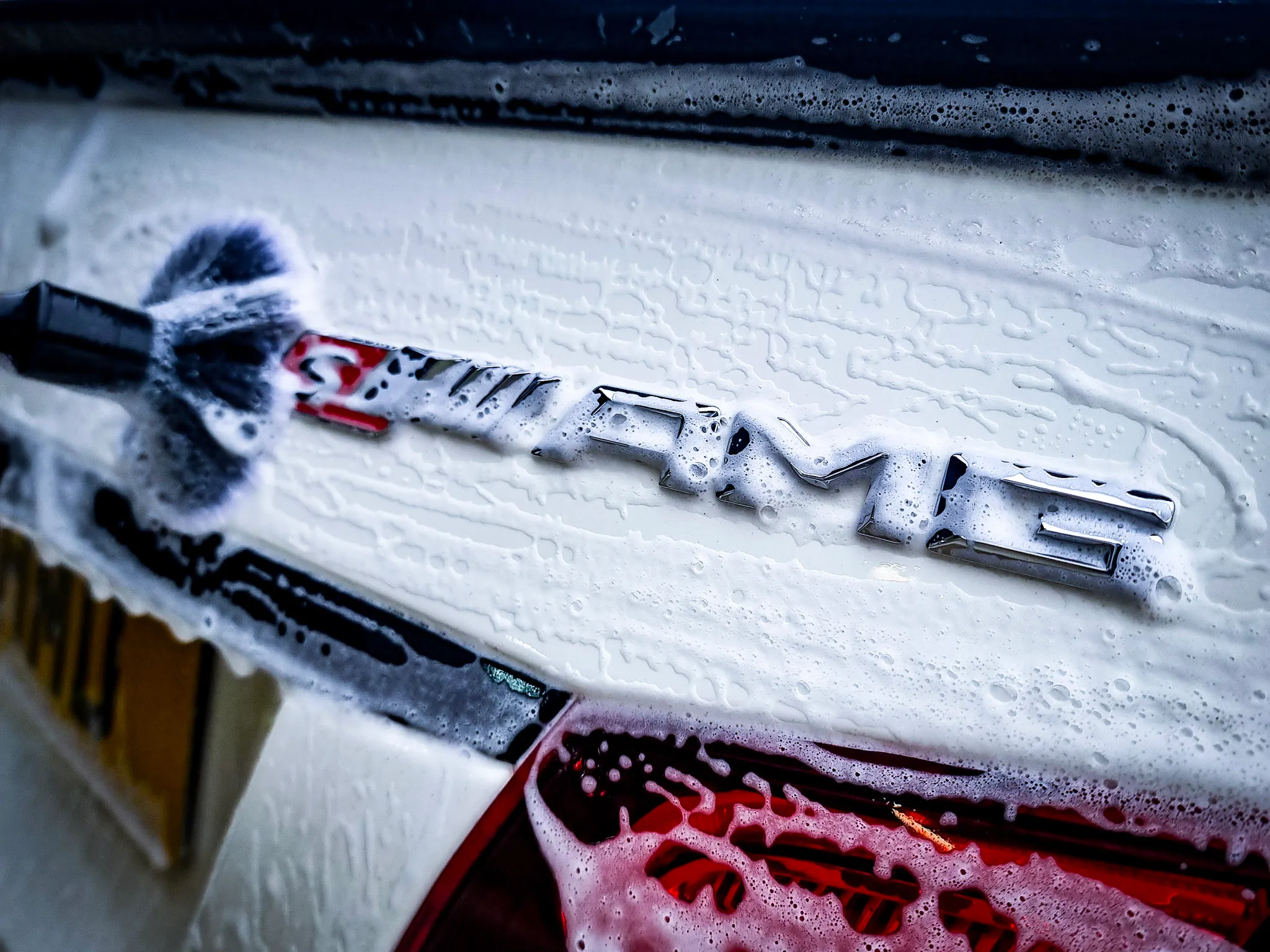 Close-up of a foam-covered Mercedes AMG badge on a white car during washing.