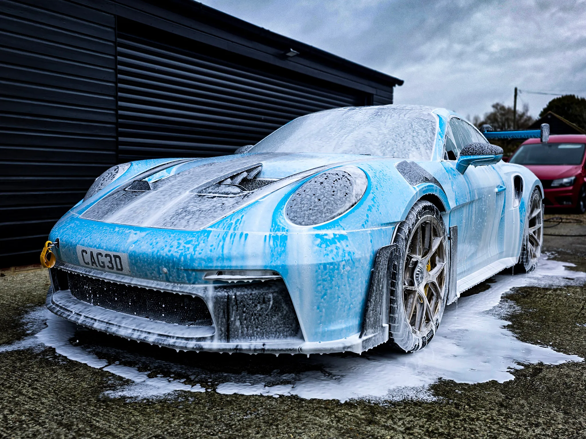 Blue sports car covered in thick white foam during a wash in front of a black building.