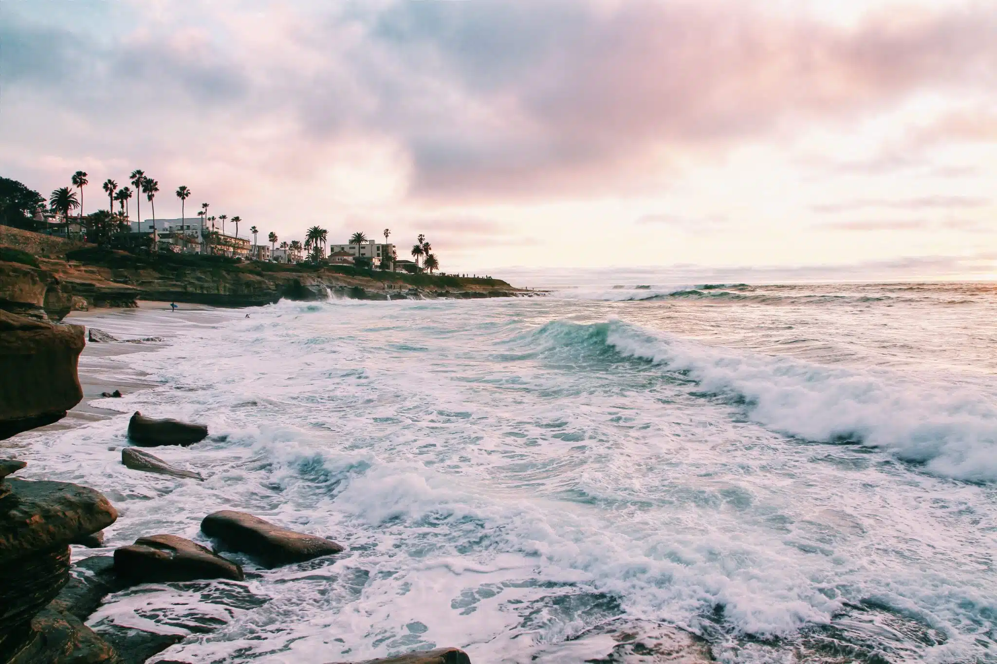 Waves crashing on a rocky beach at sunset with palm trees and houses along the shore.