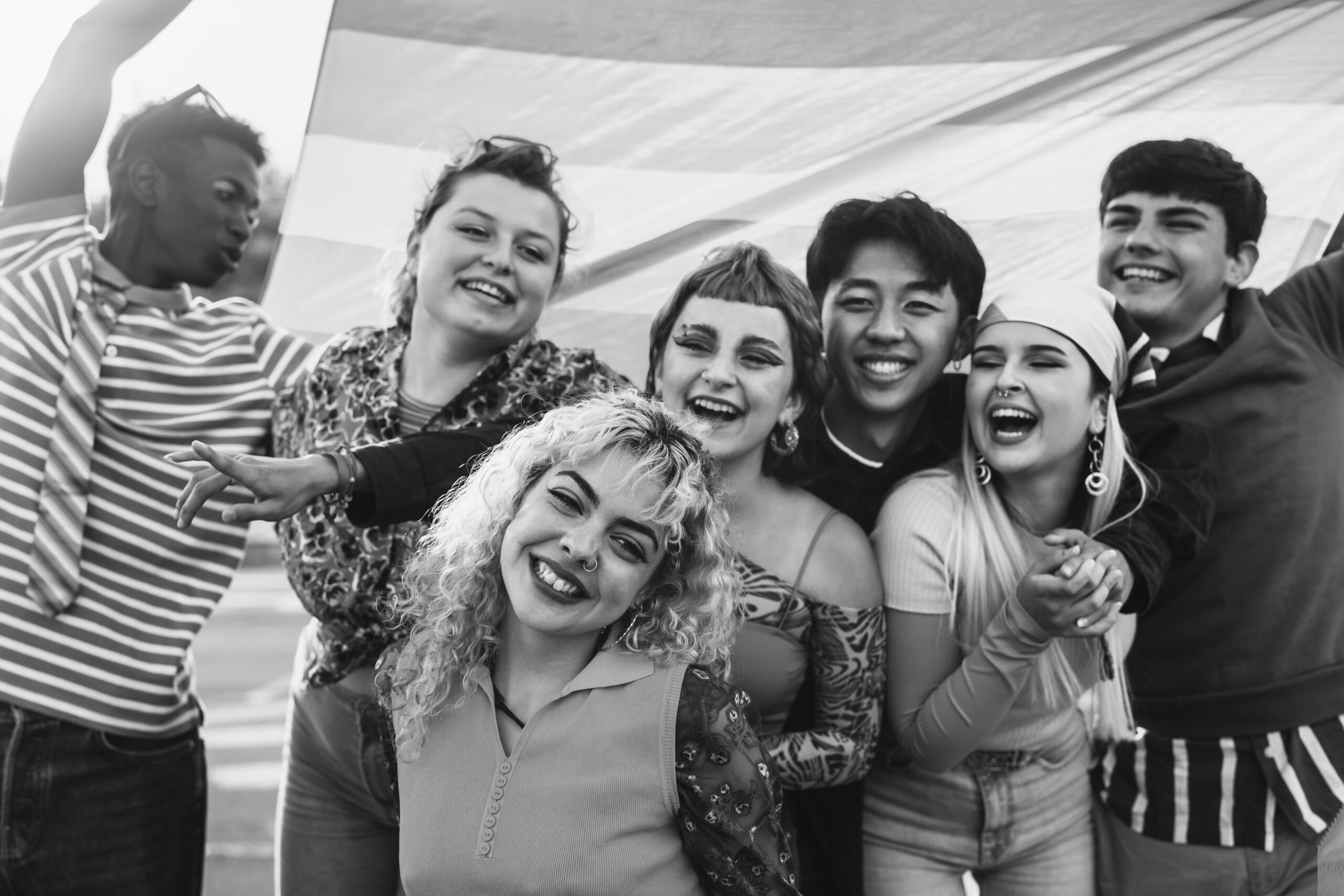 A diverse group of six young people smiling and laughing together while holding a large flag behind them.