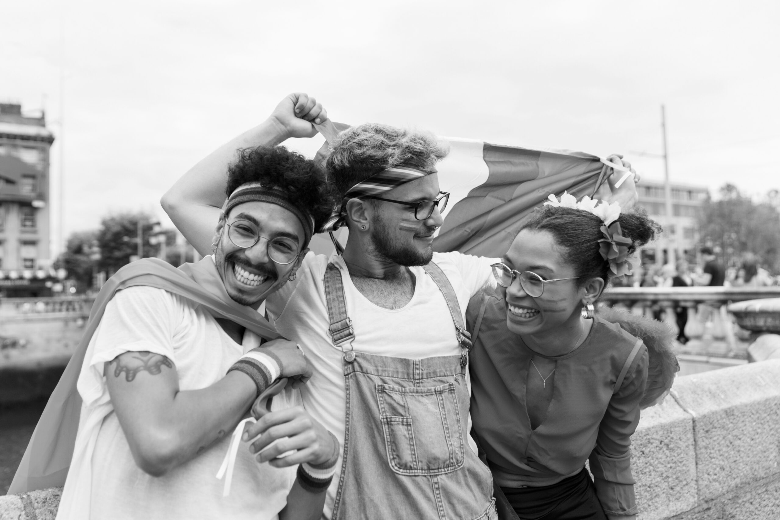 Three smiling young people with glasses and face paint celebrating outdoors with a pride flag.