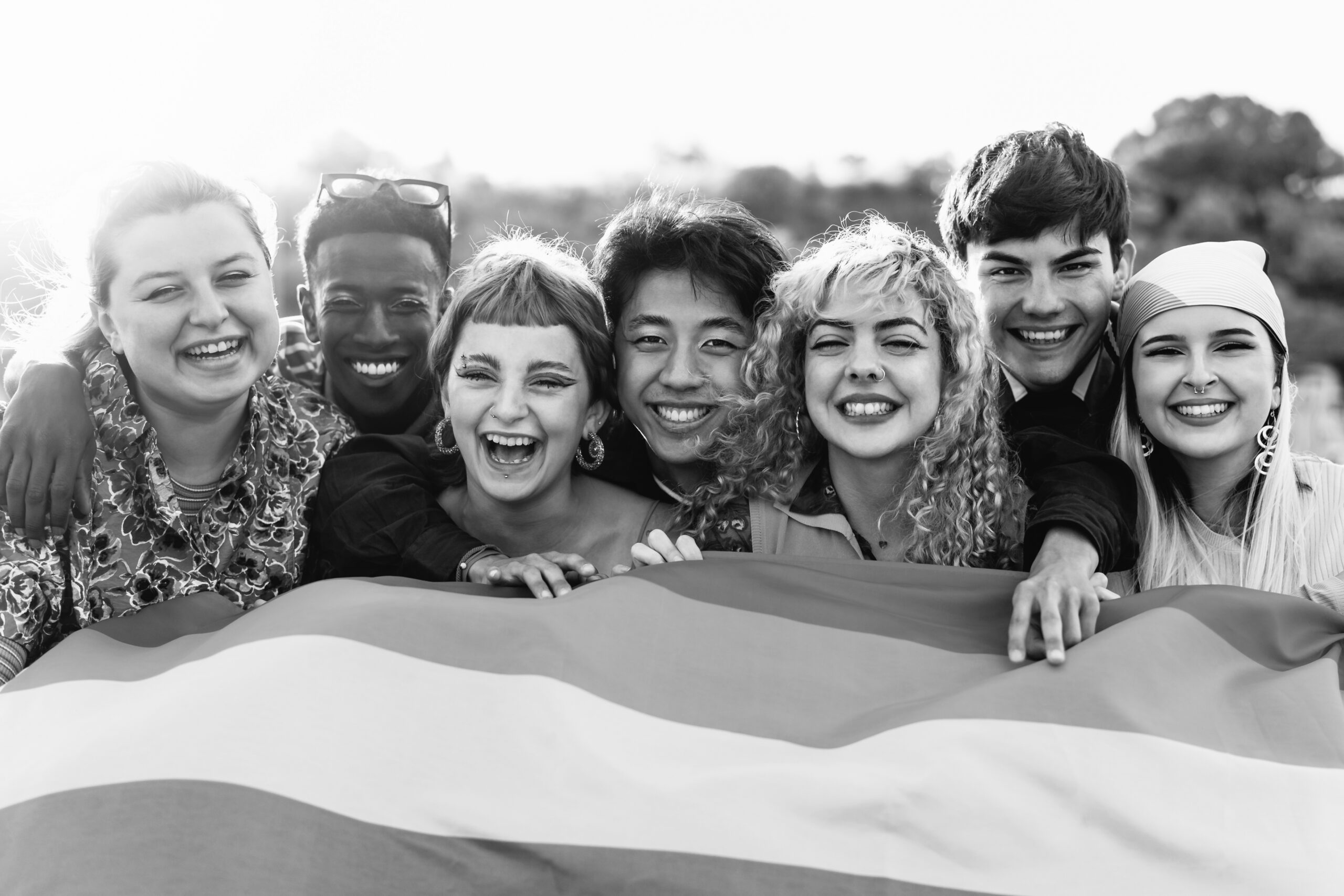 Group of seven diverse young adults smiling and holding a large pride flag outdoors.