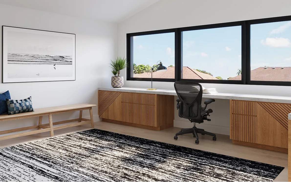 Modern home office with large windows, wooden desk, ergonomic black chair, bench with blue pillows, and a patterned black-and-white rug.