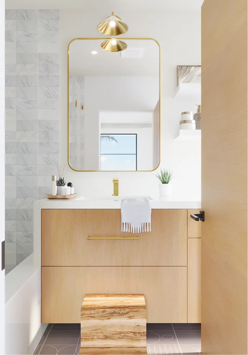 Modern bathroom vanity with light wood cabinetry, white countertop, gold faucet and framed mirror, small plant and toiletries on countertop.