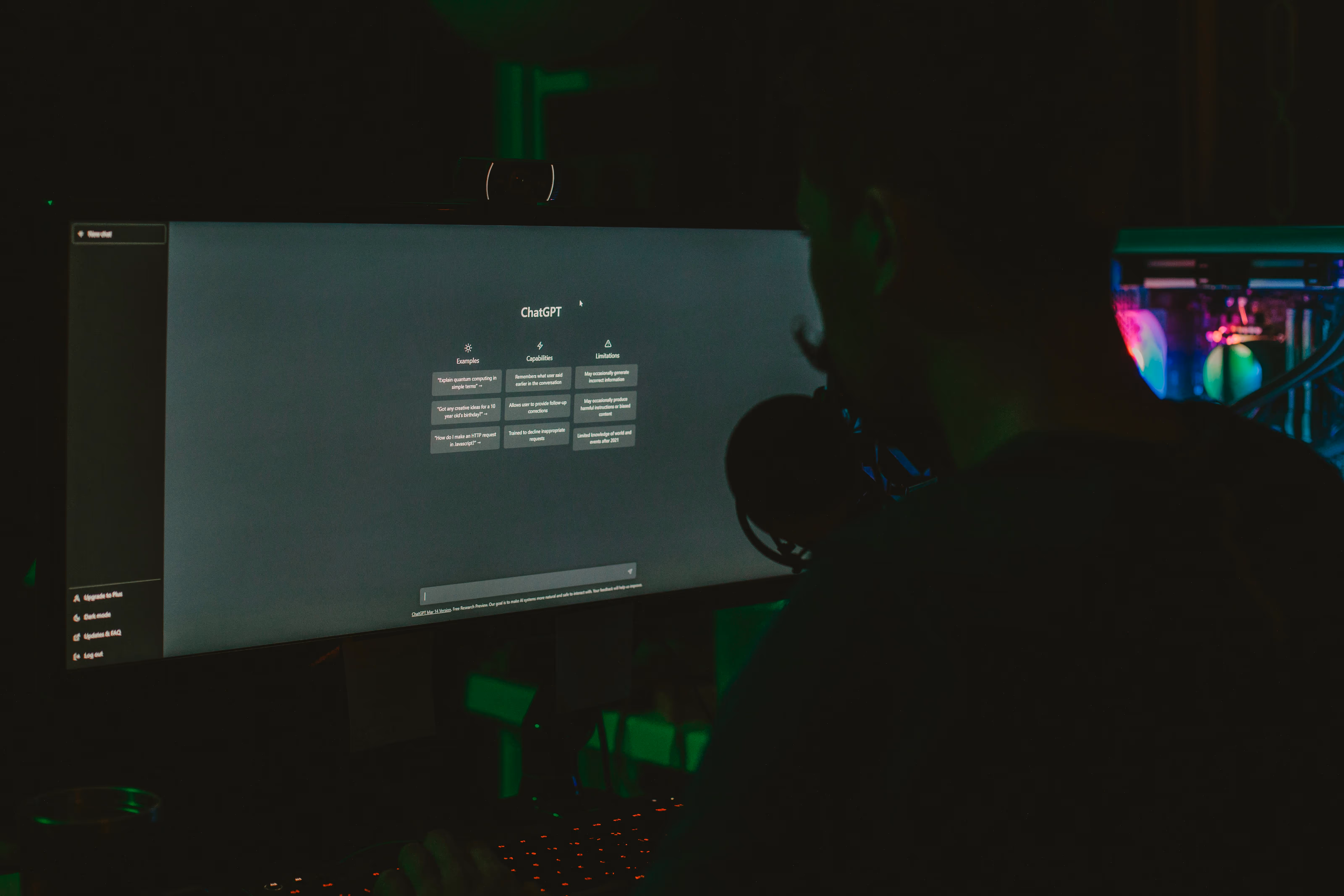 Person am Schreibtisch vor Monitor mit ChatGPT Startseite in dunklem Raum.