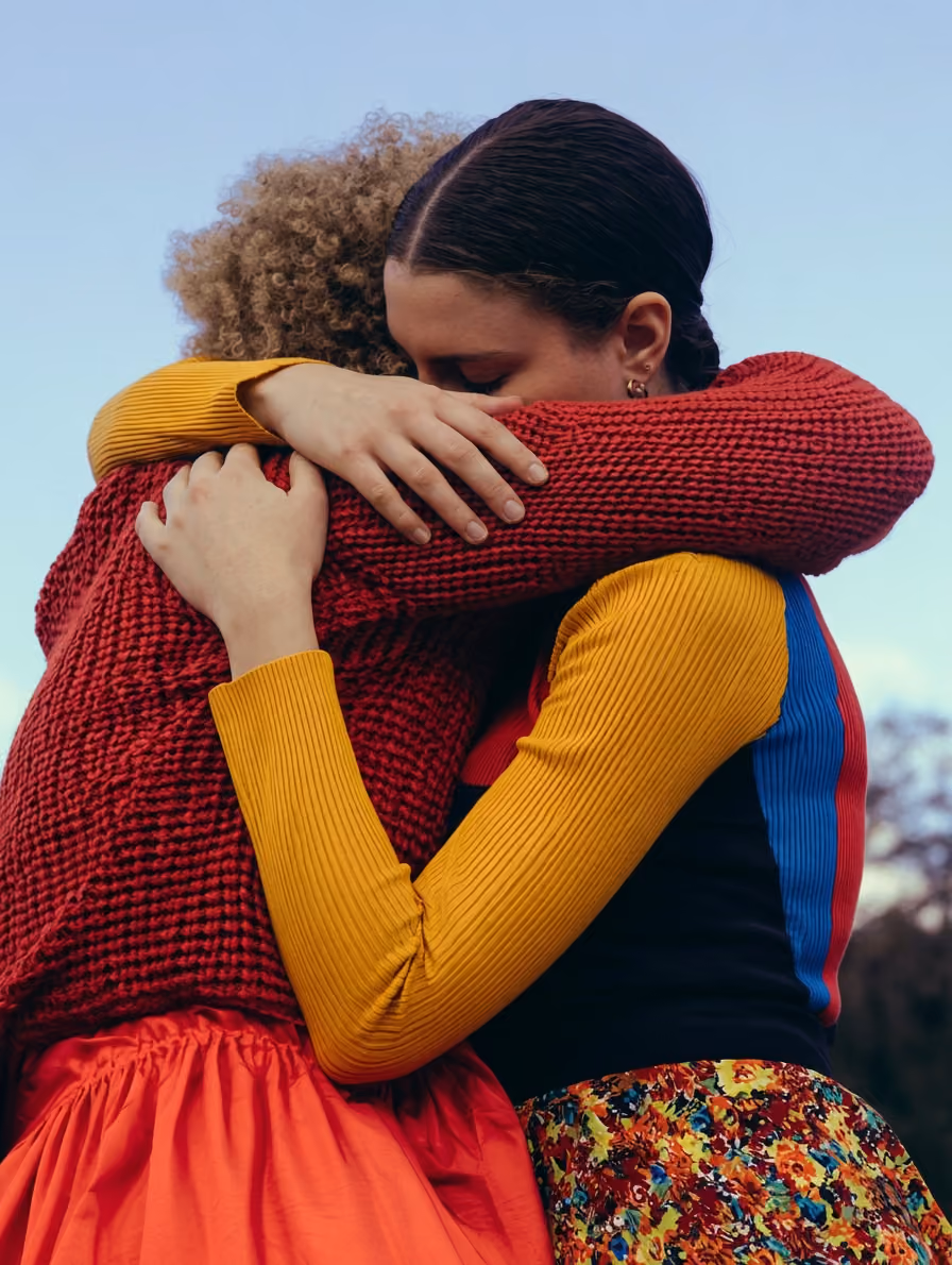Zwei Frauen umarmen sich eng, eine trägt roten Pullover, die andere gelben Ärmel mit buntem Rock.