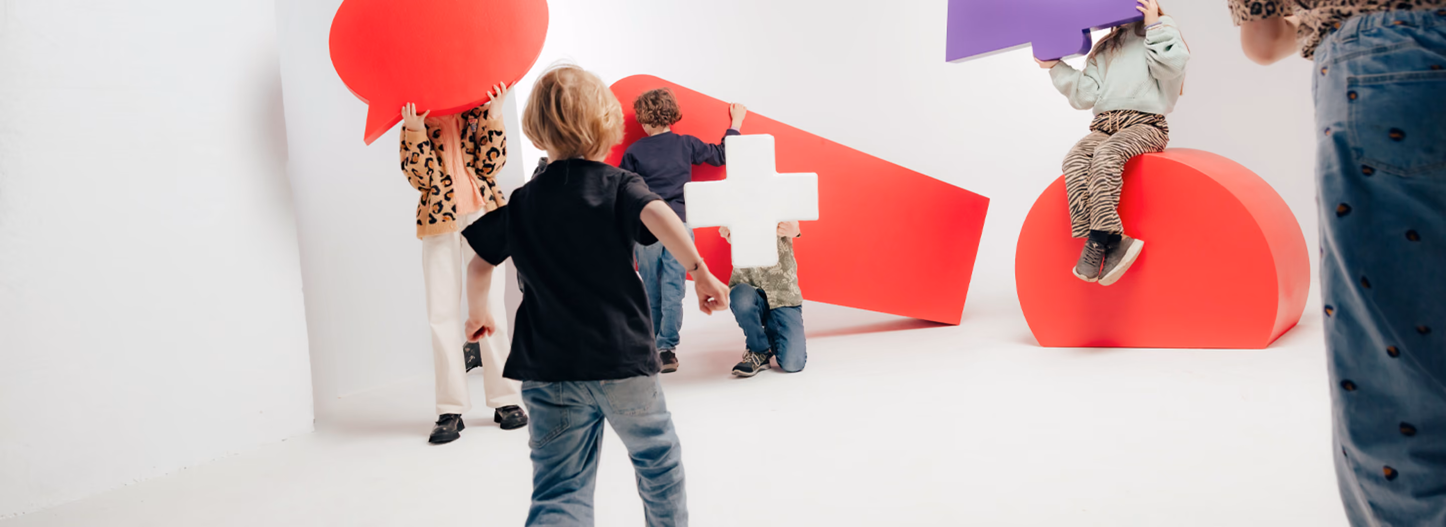 Kinder spielen mit großen roten Formen und einem weißen Pluszeichen vor weißem Hintergrund.