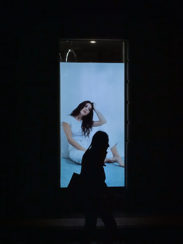 Eine Silhouette einer Frau vor einem beleuchteten Werbeplakat mit einer sitzenden Frau in weißem Kleid.